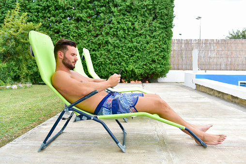 Young man on a sun lounger by the pool is using a cellphone