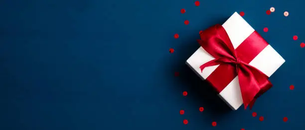 Photo of White gift box with red ribbon bow on blue background with confetti. Christmas present, valentine day surprise, birthday concept. Flat lay, top view.