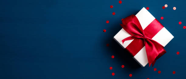 白色禮品盒，藍色背景有紅色絲帶蝴蝶結，帶紙屑。聖誕禮物，情人節驚喜，生日概念。平躺，頂視圖。 - 禮物 圖片 個照片及圖片檔