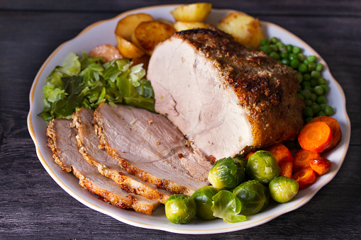 Roasted sliced ham and vegetables: potatoes, carrots, brussel sprouts, cabbage and green peas on black wooden background