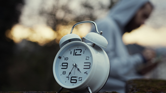 Focused on a alarm clock. Motion blur of bearded young man taking notes on notebook background