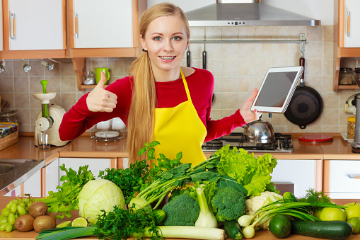 Young woman in kitchen having many green vegetables on table, holding tablet thinking about cooking something and searching for recipes in internet
