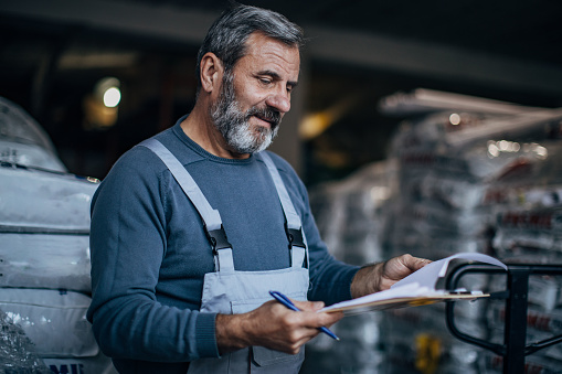 Mature man wearing jumpsuit working in the warehouse and reading the checklist