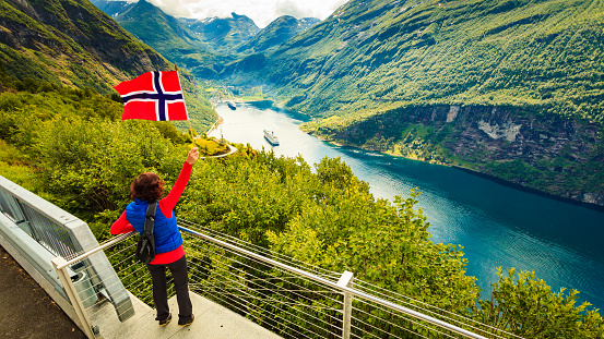 Female tourist with norwegian flag enjoying scenic view over fjord Geirangerfjord with cruise ship. Cruising vacation and travel.