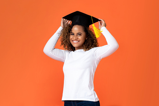 Happy smiling young African American woman wearing graduation cap isolated on orange background  for education concepts