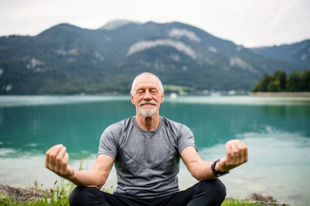 a senior man pensioner sitting by lake in nature, doing yoga exercise. - senior adult mountain hiking recreational pursuit imagens e fotografias de stock
