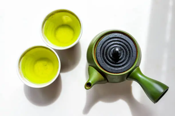 Green clay teapot with kyusu handle flat top flat lay view down on white counter background with Japanese tea brewing genmaicha or sencha in cups during ceremony