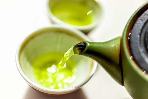 Closeup of green clay tea pot teapot on white table background and pouring liquid motion of colorful vibrant Japanese sencha or genmaicha drink during ceremony