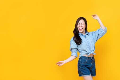 Excited pretty Asian woman smiling with arm raise isolated on yellow bakgound with copy spac