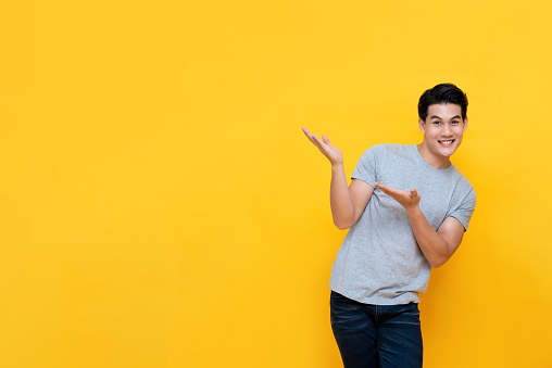 Smiling casual handsome Asian man doing presenting gesture with hands open to copy space aside isolated on yellow background