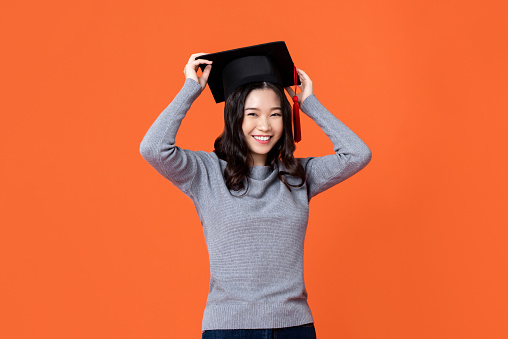 Happy smiling young Asian woman wearing graduation cap isolated on orange background for education concepts