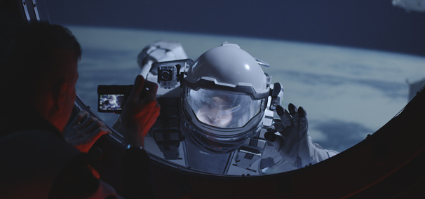 Medium close-up of a male astronaut filming his spacewalking crewmate from inside a spacecraft