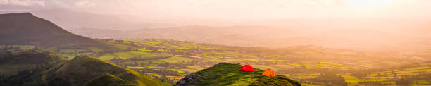 Colourful dome tents wild camping on a dramatic mountain ridge overlooking the picturesque valley below at sunset.