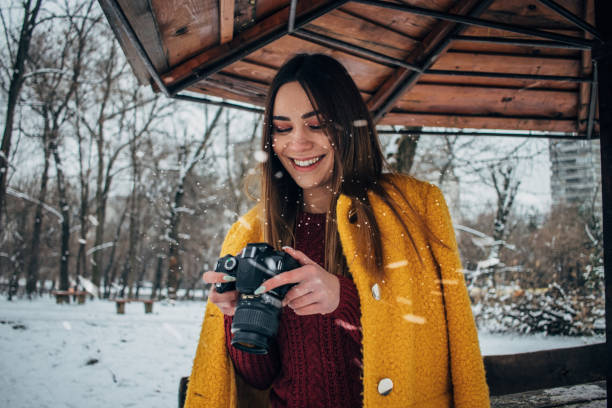Beautiful young woman in yellow coat standing in park and looking at photos on digital camera.
