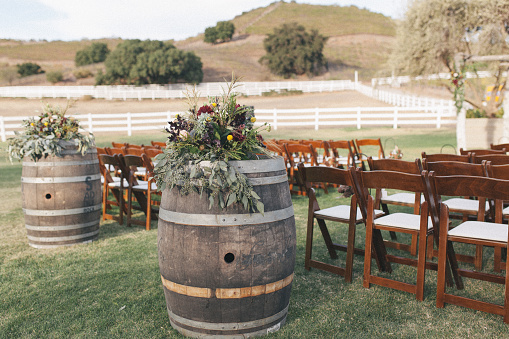 Outdoor ceremony area decorated with empty wine barrels, boho styled floral arrangement, pine cones, chairs and mountains on the background