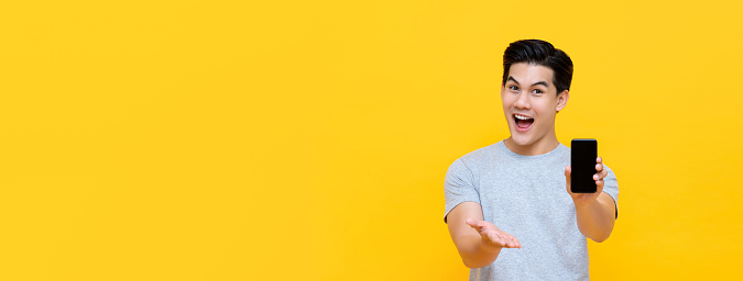 Wow excited young Asian man showing mobile phone with open hand gesture isolated on yellow banner background with copy space