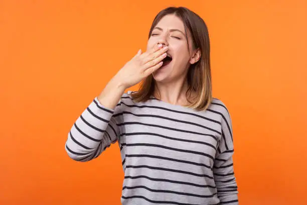 Portrait of exhausted or bored young woman with brown hair in long sleeve striped shirt standing, yawning and covering mouth with hand, disinterest. indoor studio shot isolated on orange background