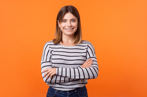 Portrait of cute happy young woman with brown hair in long sleeve shirt standing with crossed arms and looking at camera with kind toothy smile. indoor studio shot isolated on orange background