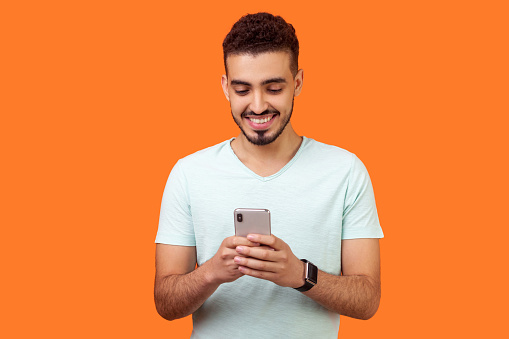 Portrait of cheerful brunette man with beard in white t-shirt using cellphone and smiling, reading good news message, enjoying mobile application. indoor studio shot isolated on orange background