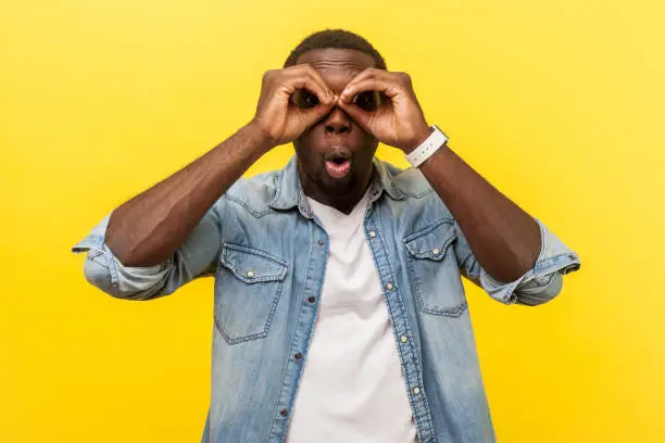Photo of Portrait of astonished young man looking at camera through fingers in binoculars gesture. indoor studio shot isolated on yellow background
