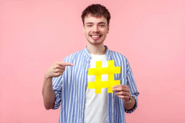 Photo of Attention to new popular hashtag. Portrait of happy brown-haired man pointing at large hash sign. isolated on pink background