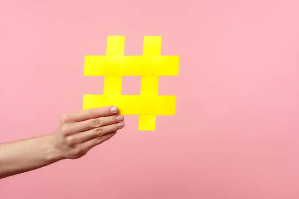 Photo of Closeup of male hand holding large yellow hashtag sign. indoor studio shot isolated on pink background