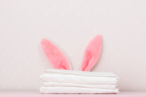 Close-up stack of clean white bedding and the Easter bunny ears, on the dressing table, on the background light walls. Copy space.