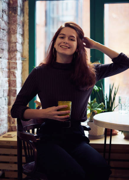 a menina esperta elegante elegante bonita bonita bonita senta-se pela janela no café, sorri belamente e bebe o smoothie ou o latte vegan saudável. mulher pensativa encantadora com cabelo marrom escuro longo - smart casual occupation casual healthy eating - fotografias e filmes do acervo