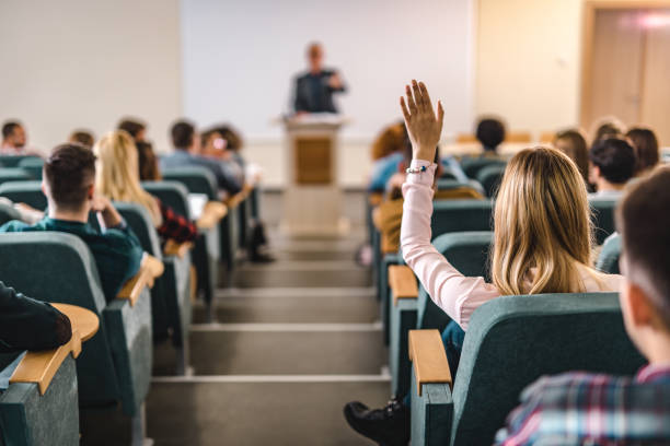 Rear view of college student raising her hand in amphitheater. Back view of female college student raising her hand to answer the question on a class at lecture hall. lecture hall photos stock pictures, royalty-free photos & images