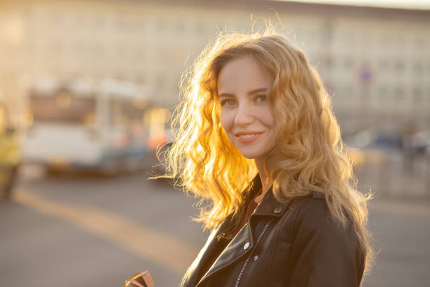 attractive curly blond girl in leather jacket standing on city street looking at camera, smiling