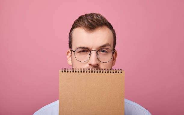 Pretty student guy in glasses covered his face with a brown loose-leaf notebook stock photo