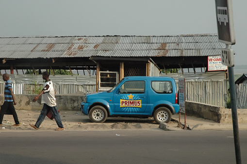 African people coming home after work, walking in front of a house on a sidewalk, passing a blue car of Primus beer at Brazzaville, Rep. du Congo on 2. February 2018, concept.