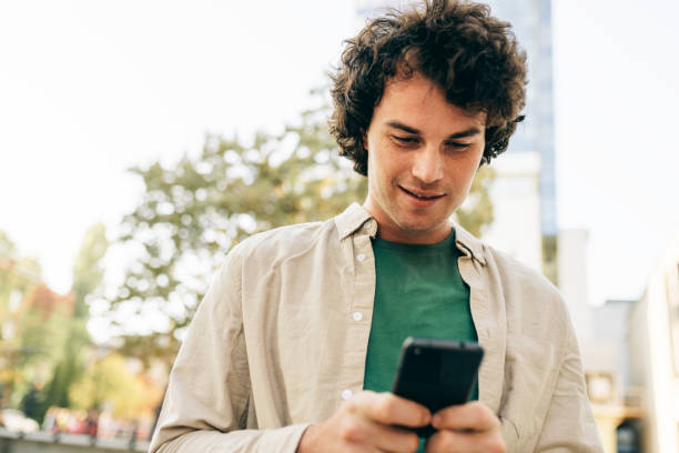istock Smart young man standing outdoors, typing messages on mobile phone. Young handsome male with curly hair browsing online on his smart phone in the city street. 1190583528