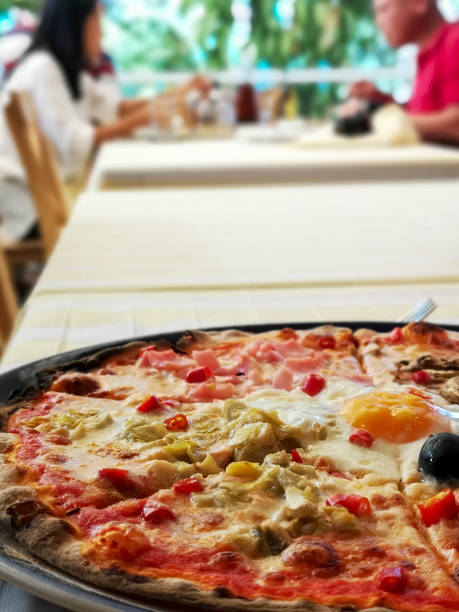 This directly above shot colorful authentic Italian crispy pizza has various toppings divided into quarters, known as four-stage, it has olives, artichoke, ham, and mushroom, along with a fresh egg and red spicy chilis, all combined and cooked in brick, wood-fired, traditional pizza oven. cuatro etapas deliciosa pizza italiana auténtica con un huevo cocinado en el medio y chiles picantes frescos, cocinado en un horno de pizza tradicional, servido en un plato, sentado en una mesa de restaurante. dos personas pueden ser vistas fuera de foco