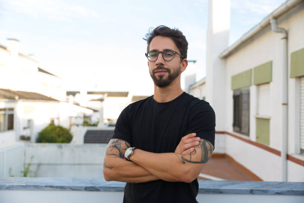 Confident stylish guy with tattoos posing on apartment balcony Confident stylish guy with tattoos posing on apartment balcony or terrace. Young man in glasses standing outside with arms crossed and looking at camera. Male portrait concept black tattoo stock pictures, royalty-free photos & images