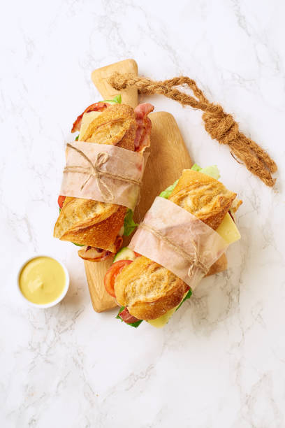 istock Two fresh big baguette sandwiches with bacon, chedder cheese, mustard, lettuce and vegetables on cutting board on marble background 1190622836