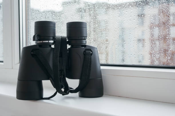 istock Black binoculars for observing neighbors is on a white window sill by the window with raindrops on the glass. Fascinating observation of the environment or nature from the window of the house 1190627946