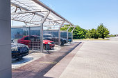 Diagonal view of the self-service car wash with four open boxes with transparent walls, in which the cars are in the process of washing. In the background green trees and blue sky