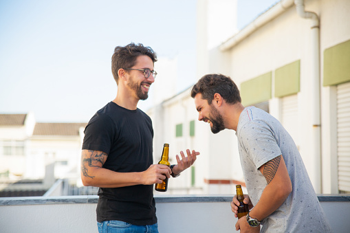 Two male friends drinking beer and sharing funny news on outdoor terrace. Two young men in casual meeting outside. Friendly chatting concept