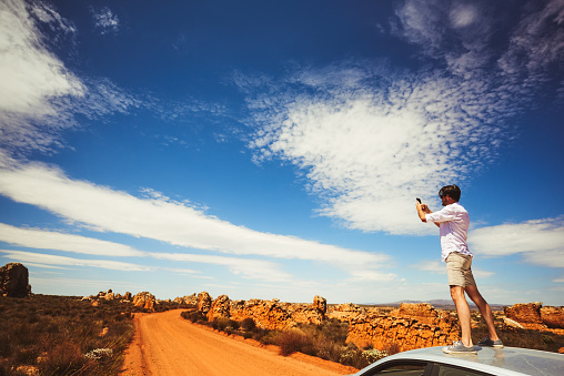 Shot of a young man using a cellphone while standing on top of a car in a rural landscape