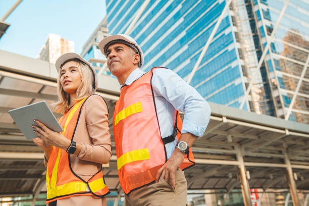 Both male and female engineers wear helmets. Standing in the construction zone while the female engineer was holding the tablet against building ,city view.