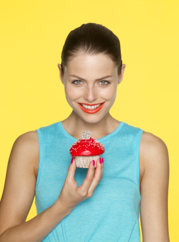 yellow background, young female, tanned, blue vest, brown hair, red painted lips, iced cake, red icing, sprinkles, marshmallow, sponge cake, smile