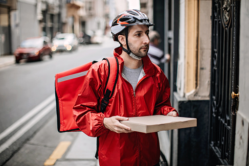 Delivery boy in red uniform holding  pizza box in front of door, making a home delivery