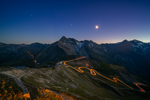 light trails on curvy high alpine road through european mountains at night with star shapped moon
