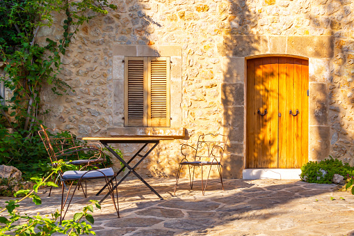 The facade of an old stone house with wooden brown shutters and door. Old armchairs and table. Majorca. Spain.