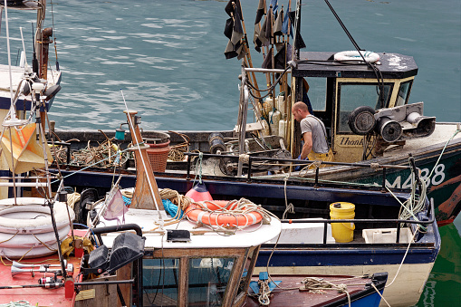 Fisherman in oilskin trousers working on his boat among ropes, nets and other fishing gear. Moored to another boat that is moored to quayside