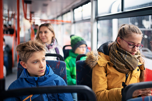 Kids travelling to school by bus or tram. Girl is checking her smartphone. Her brothers are looking out of window. Autumn time, kids are wearing warm clothing.\nNikon D850