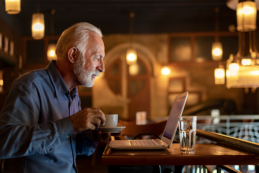 Handsome Senior Businessman with Grey Hair and Beard is Having a Coffee Break in City Café. He is Sitting at the Table in Coffee Shop and Enjoying in Hot Cup of Coffee or Tea. Older Man is Using his Laptop and Relaxing in Bar in the City Center.