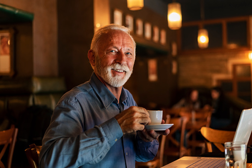 Handsome Senior Businessman with Grey Hair and Beard is Having a Coffee Break in City Café. He is Sitting at the Table in Coffee Shop and Enjoying in Hot Cup of Coffee or Tea. Older Man is Using his Laptop and Relaxing in Bar in the City Center.