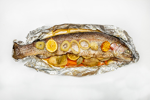 An oven roasted trout in a aluminium foil on a whitebackground. With carrots,lemon slice and onions ringon the side.